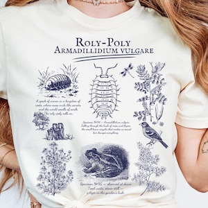 May include: Cream-colored t-shirt featuring a detailed blue illustration of a Roly-Poly, also known as Armadillidium Vulgare, with scientific drawings of insects, plants, and a frog. The text "ROLY-POLY ARMADILLIDIUM VULGARE" is printed at the top.