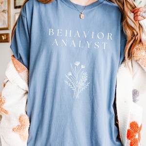 May include: A light blue t-shirt with the text "BEHAVIOR ANALYST" in white lettering and a white floral design.