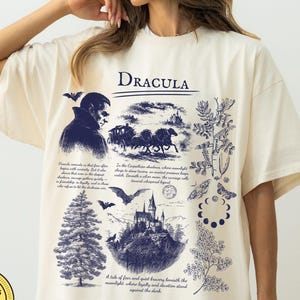 May include: Ivory-coloured t-shirt featuring a dark blue Dracula graphic. The graphic includes illustrations of a vampire, a horse-drawn carriage, a castle, bats, and text. The shirt is made of Comfort Colors material.