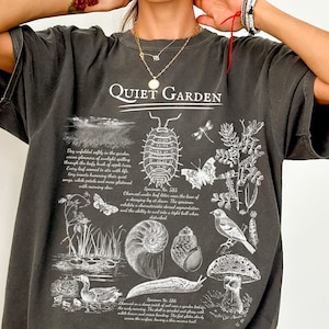 May include: Dark gray t-shirt with the text "Quiet Garden" and detailed illustrations of insects, plants, and a bird. The design includes scientific-style drawings of a pill bug, butterflies, a snail, and a mushroom.