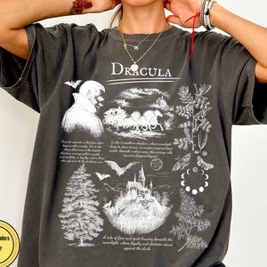 Op de afbeelding: Donkergrijs t-shirt met een grafisch ontwerp met het woord "Dracula". Het ontwerp bevat illustraties van een vampier, een paardenkoets, een kasteel, vleermuizen en botanische elementen. Het shirt is gelabeld "Comfort Colors Pepper".
