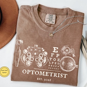 May include: Brown t-shirt with white Optometrist graphic, eye chart, and eye diagrams. The shirt has the text "OPTOMETRIST EST. 2026" and a Comfort Colors label. A silver necklace with a pendant is on the shirt.