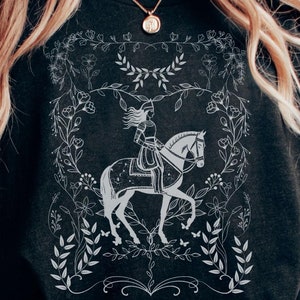 May include: A black t-shirt with a white graphic design of a woman riding a horse in a floral wreath. The design is detailed and intricate, with leaves, flowers, and butterflies.