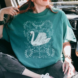 K&ouml;nnte beinhalten: Ein t&uuml;rkisfarbenes T-Shirt mit einem wei&szlig;en Schwanen-Motiv, umgeben von floralen Designs. Der Schwan ist in einem stilisierten, fast Art-Deco-Stil gehalten.
