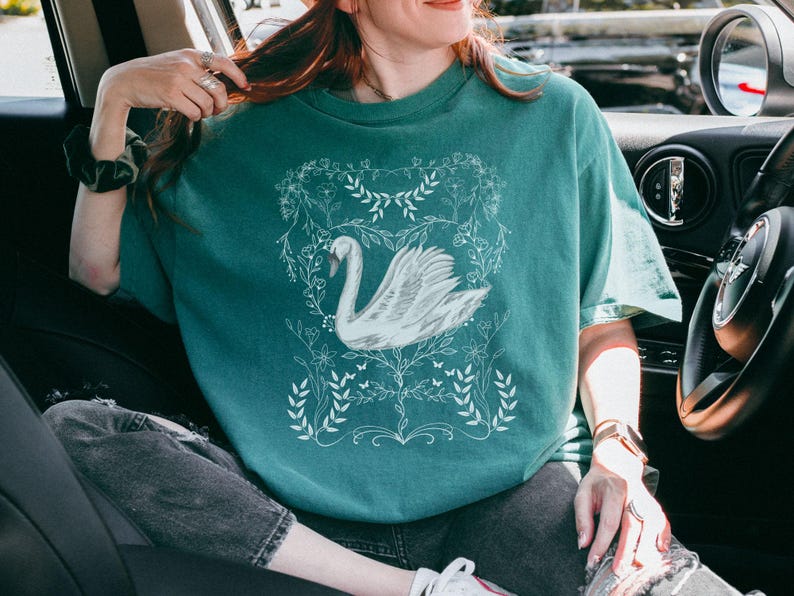 K&ouml;nnte beinhalten: Ein t&uuml;rkisfarbenes T-Shirt mit einem wei&szlig;en Schwanen-Motiv, umgeben von floralen Designs. Der Schwan ist in einem stilisierten, fast Art-Deco-Stil gehalten.