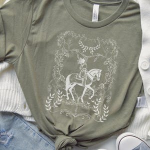May include: A green t-shirt with a white design of a woman riding a horse in a floral wreath. The design is reminiscent of a fairytale illustration.