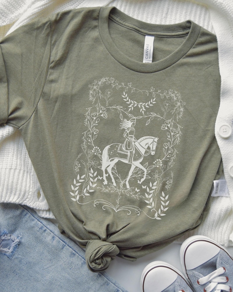 May include: A green t-shirt with a white design of a woman riding a horse in a floral wreath. The design is reminiscent of a fairytale illustration.