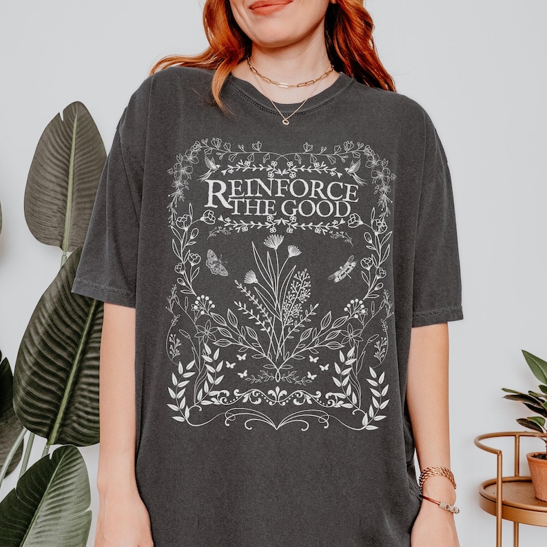 May include: A dark gray oversized t-shirt with a white graphic design featuring a floral border and the text "Reinforce the Good".