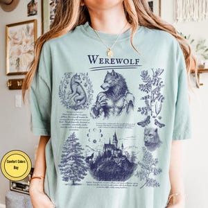 May include: A light green t-shirt featuring a dark blue graphic print with the text "WEREWOLF" at the top. The design includes illustrations of a werewolf, a wolf, a castle, a pine tree, moon phases, and botanical elements with descriptive text.