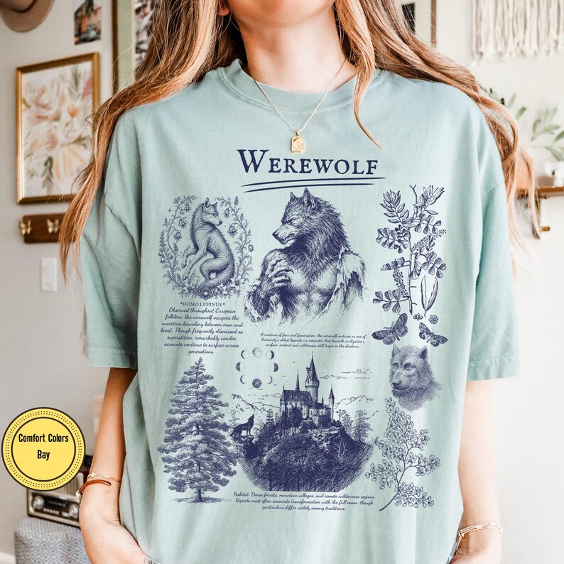 May include: A light green t-shirt featuring a dark blue graphic print with the text "WEREWOLF" at the top. The design includes illustrations of a werewolf, a wolf, a castle, a pine tree, moon phases, and botanical elements with descriptive text.