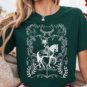 May include: A green t-shirt with a white design of a woman riding a horse surrounded by floral details.