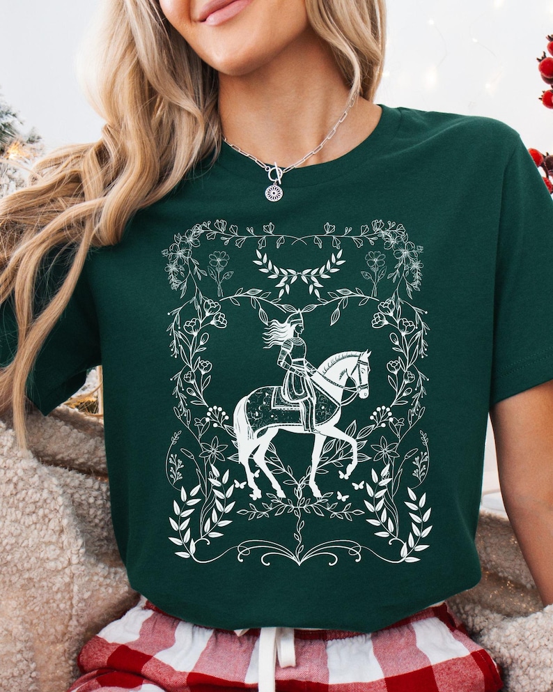 May include: A green t-shirt with a white design of a woman riding a horse surrounded by floral details.