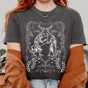 May include: A dark gray t-shirt with a white graphic design of a wolf and a knight fighting inside a heart-shaped floral frame.