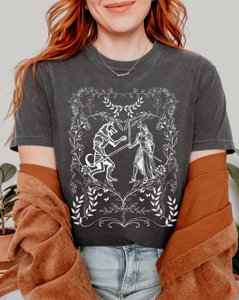 May include: A dark gray t-shirt with a white graphic design of a wolf and a knight fighting inside a heart-shaped floral frame.