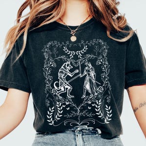 May include: A black t-shirt with a white graphic design of a wolf and a knight fighting. The design is surrounded by a floral border.