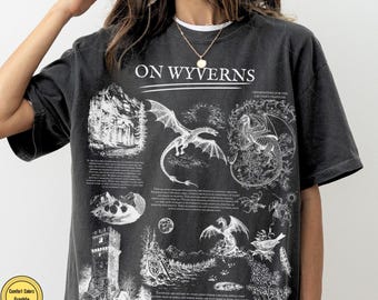 May include: A charcoal gray short-sleeve t-shirt featuring a white graphic print titled ON WYVERNS. The design includes detailed illustrations of wyverns, a stone tower, mountain landscapes, and small blocks of text describing the creatures.