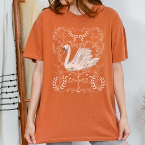 K&ouml;nnte beinhalten: Ein orangefarbenes T-Shirt mit einem wei&szlig;en Schwanen-Motiv und floralem Design. Der Schwan befindet sich in einem zarten, floralen Rahmen.