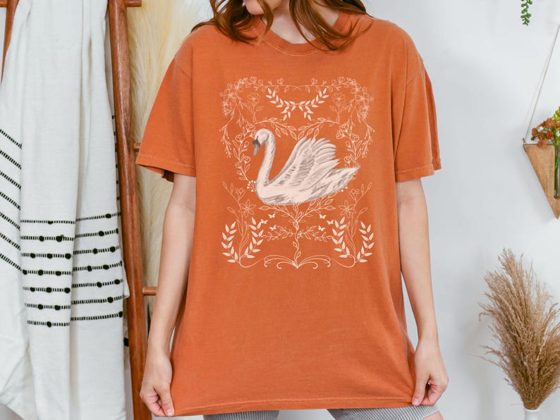 K&ouml;nnte beinhalten: Ein orangefarbenes T-Shirt mit einem wei&szlig;en Schwanen-Motiv und floralem Design. Der Schwan befindet sich in einem zarten, floralen Rahmen.