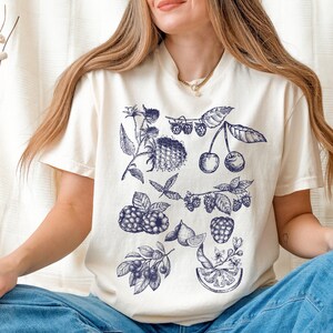 May include: A white t-shirt with a blue and white illustration of various fruits and berries, including cherries, raspberries, and blueberries.