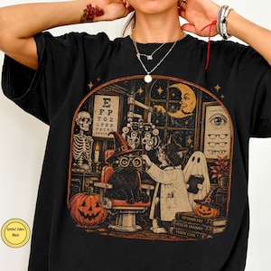 May include: A black t-shirt featuring a graphic print of a cat wearing a witch hat and glasses sitting in an optometry chair. The scene includes a skeleton, a ghost, a crescent moon, jack-o'-lanterns, and books titled Optometry, Ocular Anatomy, and Vision Care.