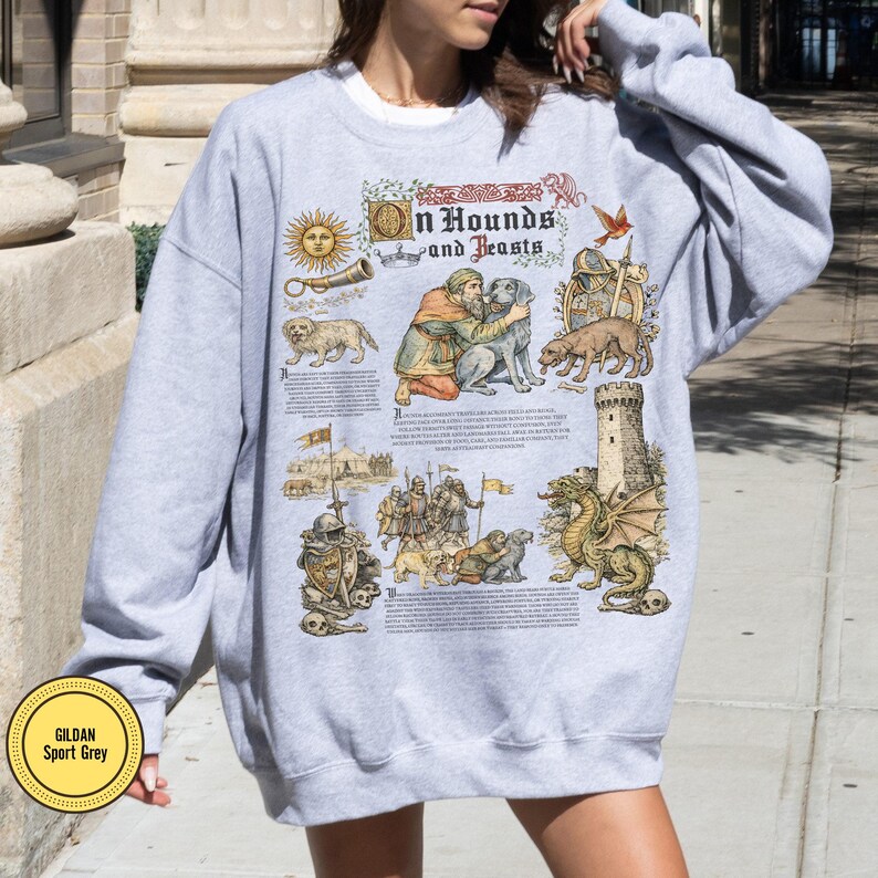 May include: Light gray Gildan Sport Grey sweatshirt featuring a medieval-style illustration with the text "On Hounds and Beasts." The design includes dogs, a dragon, and figures in historical attire. The sweatshirt is a crew neck.