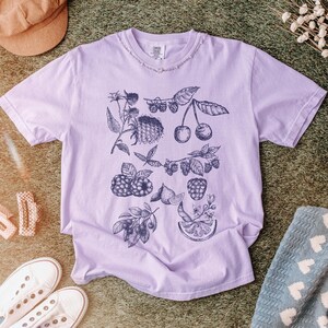 May include: A lavender t-shirt with a black and white illustration of various fruits and berries, including cherries, raspberries, and lemons.