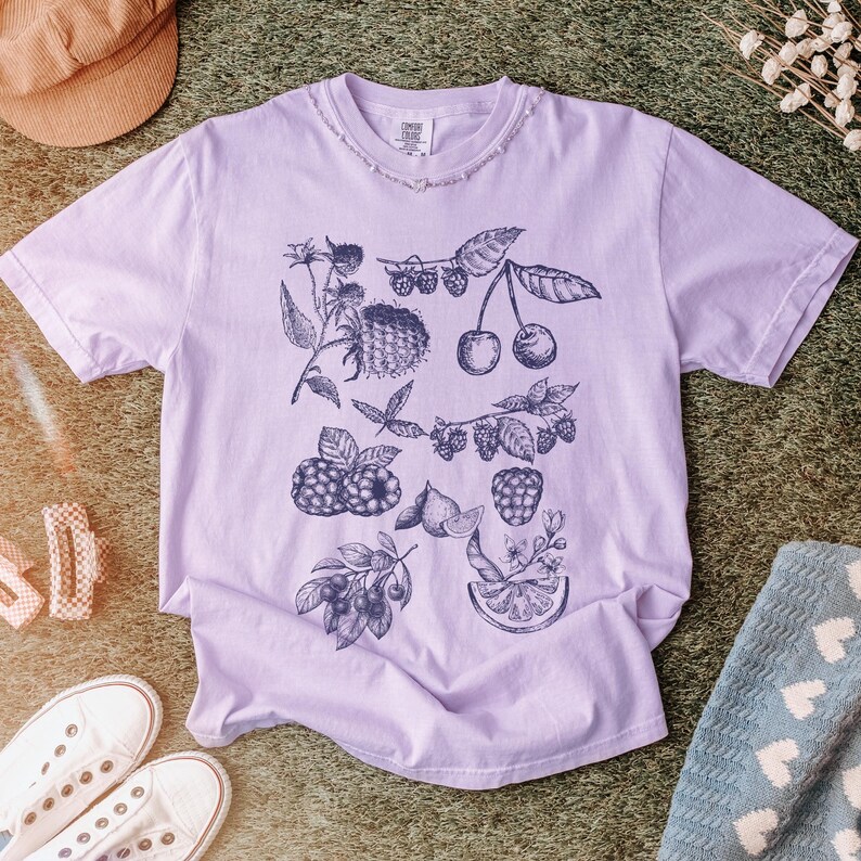 May include: A lavender t-shirt with a black and white illustration of various fruits and berries, including cherries, raspberries, and lemons.