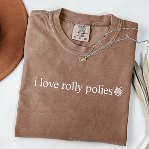 May include: A brown Comfort Colors t-shirt with the phrase "i love rolly polies" in white text. A small insect graphic is next to the text. A silver necklace with a pendant is draped across the shirt.