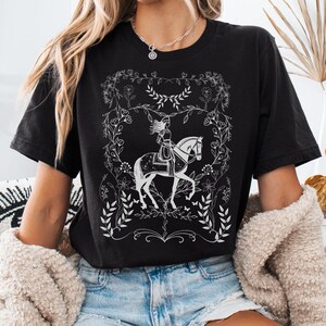 May include: Black t-shirt with a white graphic of a woman riding a horse in a floral frame.