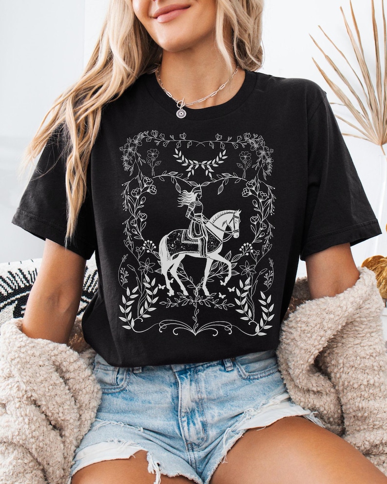 May include: Black t-shirt with a white graphic of a woman riding a horse in a floral frame.