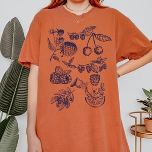May include: A burnt orange t-shirt with a blue and white graphic print of various fruits and berries, including cherries, raspberries, and lemons.