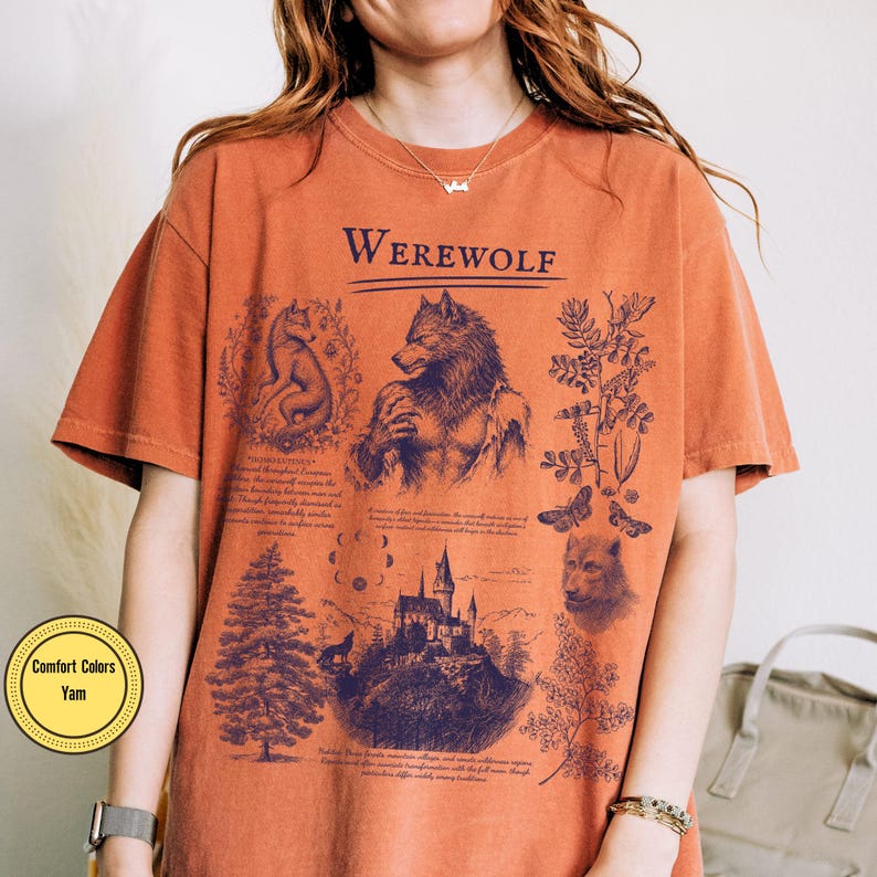 May include: A burnt orange short-sleeve t-shirt featuring a dark blue graphic print with the text Werewolf. The design includes illustrations of a wolf, a castle, a pine tree, moon phases, and botanical elements with small blocks of text.
