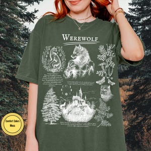 May include: A moss green short-sleeve t-shirt featuring a white graphic print with the text "WEREWOLF" at the top. The design includes illustrations of a werewolf, a wolf, a castle, moon phases, pine trees, and botanical elements with text.