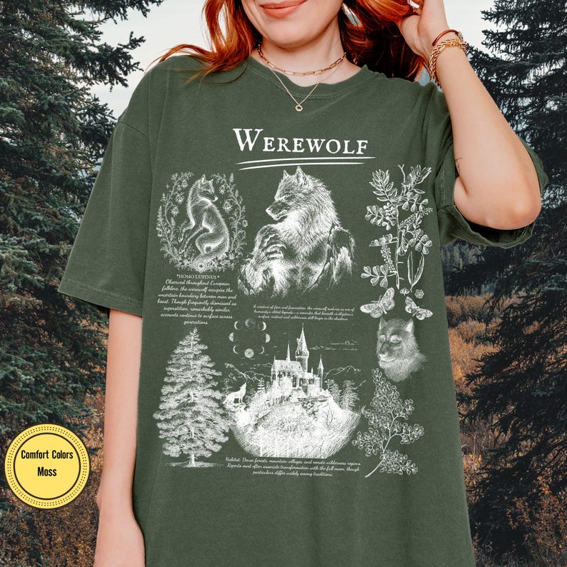 May include: A moss green short-sleeve t-shirt featuring a white graphic print with the text "WEREWOLF" at the top. The design includes illustrations of a werewolf, a wolf, a castle, moon phases, pine trees, and botanical elements with text.