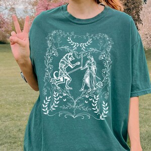 May include: A green t-shirt with a white graphic print of a wolf and a knight fighting in a heart-shaped frame surrounded by floral designs.