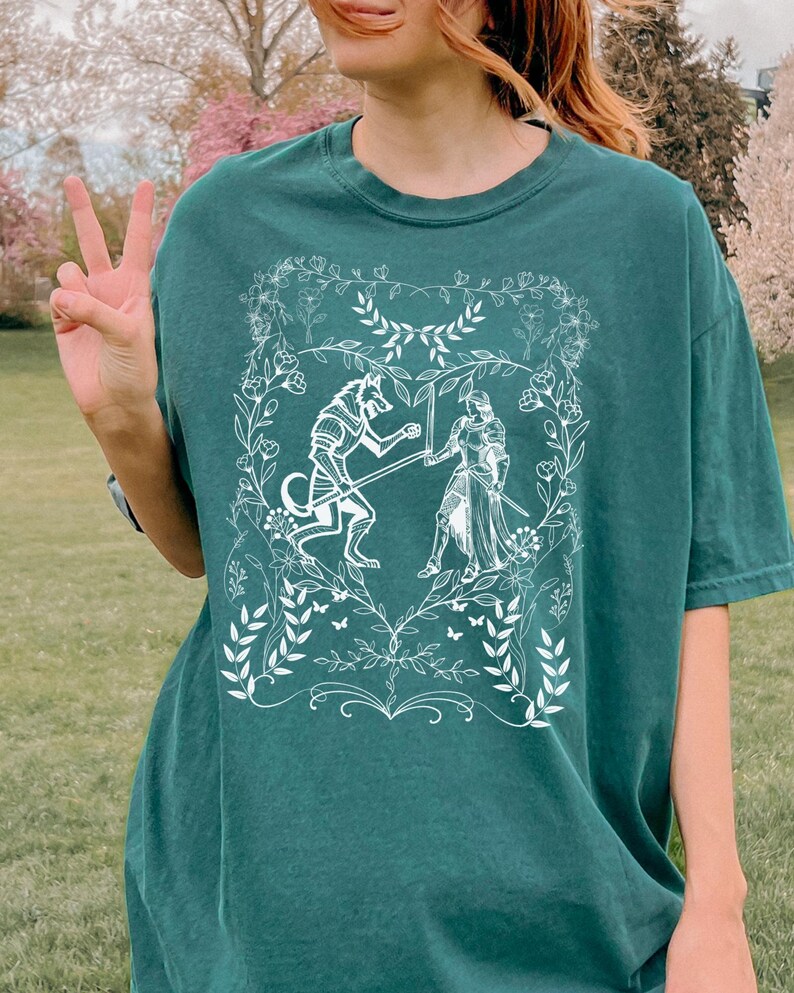 May include: A green t-shirt with a white graphic print of a wolf and a knight fighting in a heart-shaped frame surrounded by floral designs.