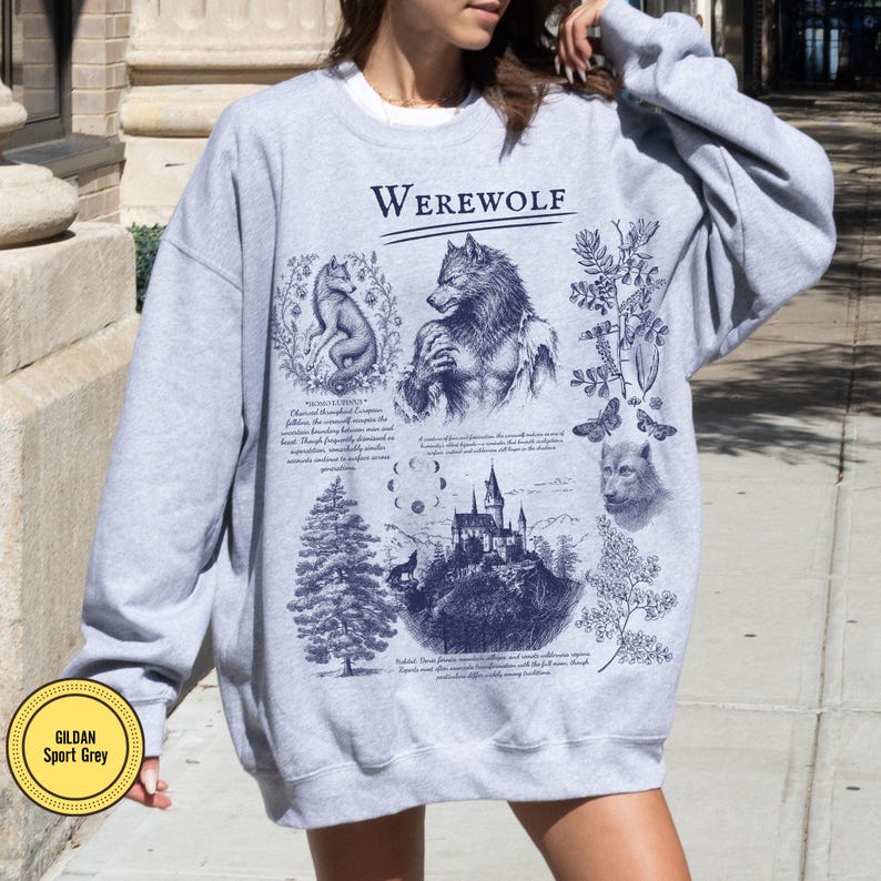 May include: A light gray crewneck sweatshirt featuring a dark blue graphic print with the text "WEREWOLF" at the top. The design includes illustrations of a wolf, a werewolf, a castle, a moon cycle, pine trees, and botanical elements.