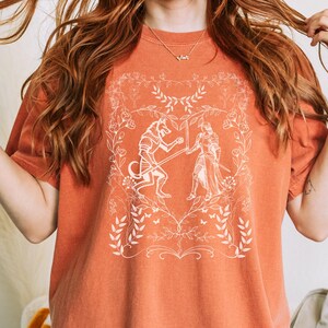 May include: A burnt orange t-shirt with a white graphic design of a knight fighting a wolf in a floral frame.