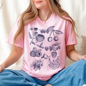 May include: A pink t-shirt with a blue and white graphic print of various fruits and berries, including cherries, raspberries, and blueberries.