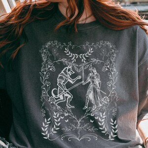 May include: A dark gray t-shirt with a white graphic design of a wolf and a knight fighting. The design is surrounded by a floral border.