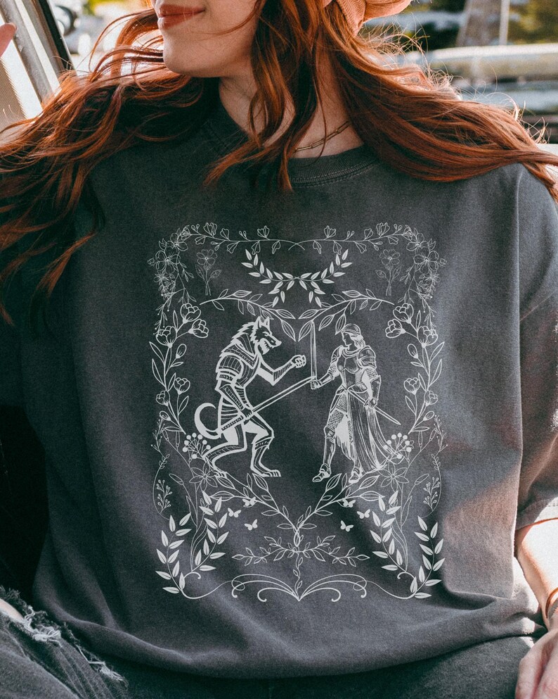 May include: A dark gray t-shirt with a white graphic design of a wolf and a knight fighting. The design is surrounded by a floral border.