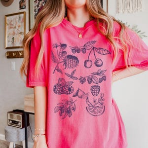 May include: A pink t-shirt with a blue graphic print of various fruits and berries, including cherries, raspberries, and lemons.