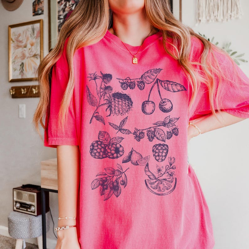 May include: A pink t-shirt with a blue graphic print of various fruits and berries, including cherries, raspberries, and lemons.