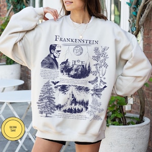 May include: Cream-colored sweatshirt with a dark blue graphic featuring the word "Frankenstein" above an illustration of the monster, a wolf, a full moon, and other gothic imagery. The sweatshirt has a round neckline and long sleeves.