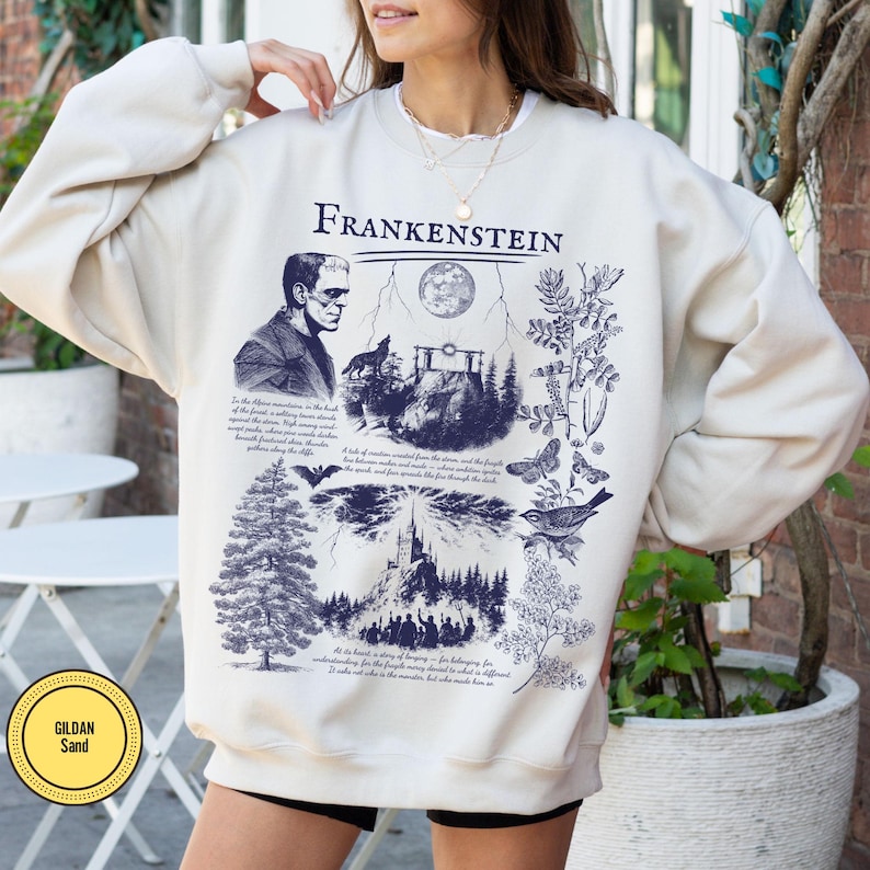May include: Cream-colored sweatshirt with a dark blue graphic featuring the word "Frankenstein" above an illustration of the monster, a wolf, a full moon, and other gothic imagery. The sweatshirt has a round neckline and long sleeves.