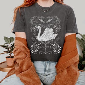 K&ouml;nnte beinhalten: Ein dunkelgraues T-Shirt mit einem wei&szlig;en Motiv eines Schwans in einem Blumenkranz. Der Schwan ist nach rechts gerichtet und hat einen langen Hals. Der Blumenkranz besteht aus Bl&auml;ttern und Blumen. Das T-Shirt wird von einer Person mit roten Haaren und einer braunen Jacke getragen.