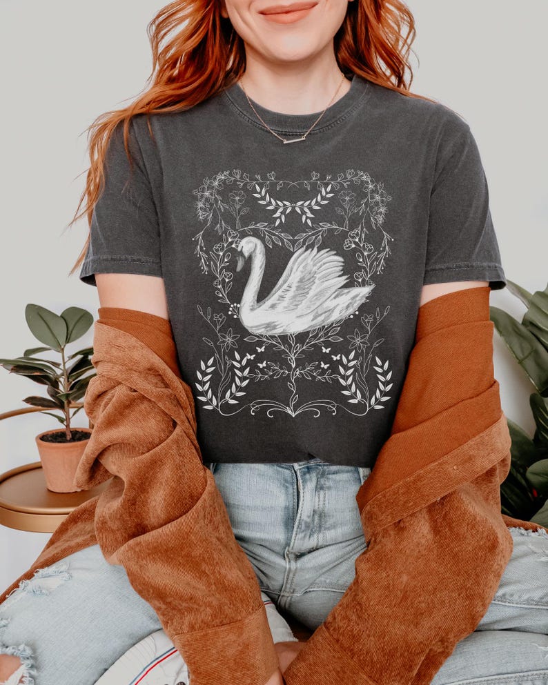 K&ouml;nnte beinhalten: Ein dunkelgraues T-Shirt mit einem wei&szlig;en Motiv eines Schwans in einem Blumenkranz. Der Schwan ist nach rechts gerichtet und hat einen langen Hals. Der Blumenkranz besteht aus Bl&auml;ttern und Blumen. Das T-Shirt wird von einer Person mit roten Haaren und einer braunen Jacke getragen.