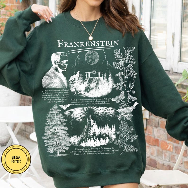 May include: Dark green sweatshirt with a white graphic design featuring the word "Frankenstein" at the top. The design includes an illustration of Frankenstein's monster, a wolf, a full moon, a castle, and botanical elements. The sweatshirt has a crew neck.