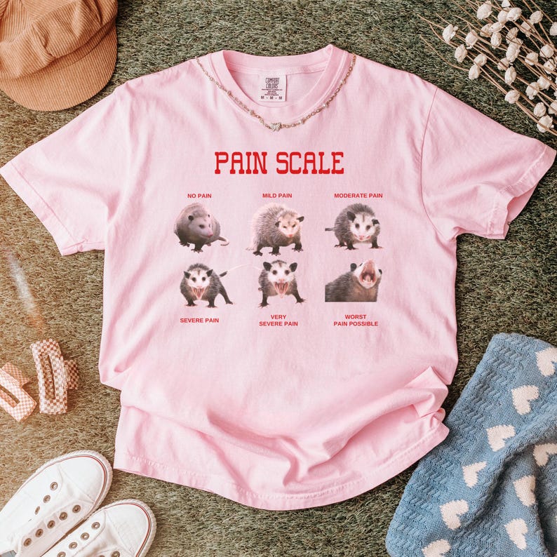 May include: Pink t-shirt with a pain scale chart featuring images of opossums. The chart reads "Pain Scale" with the following levels: "No Pain", "Mild Pain", "Moderate Pain", "Severe Pain", "Very Severe Pain", and "Worst Pain Possible".