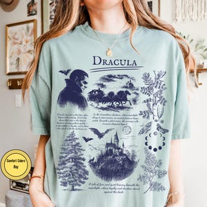 Op de afbeelding: Lichtgroen T-shirt met een donkerblauw grafisch ontwerp met het woord "DRACULA". Het ontwerp bevat illustraties van Dracula, een koets, een kasteel, vleermuizen en botanische elementen. Tekst is ook inbegrepen.